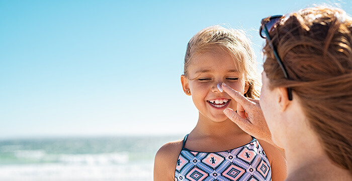 Fillette blonde en maillot de bain à qui la mère applique de la crème solaire sur le visage