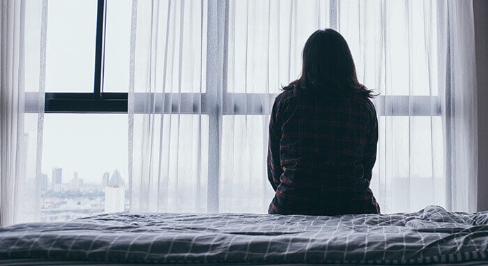 femme de dos, assisie sur un lit, regardant tristement par la fenêtre