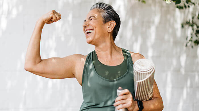 femme d'un certain âge contractant son biceps en riant avec un tapis de yoga sous le bras