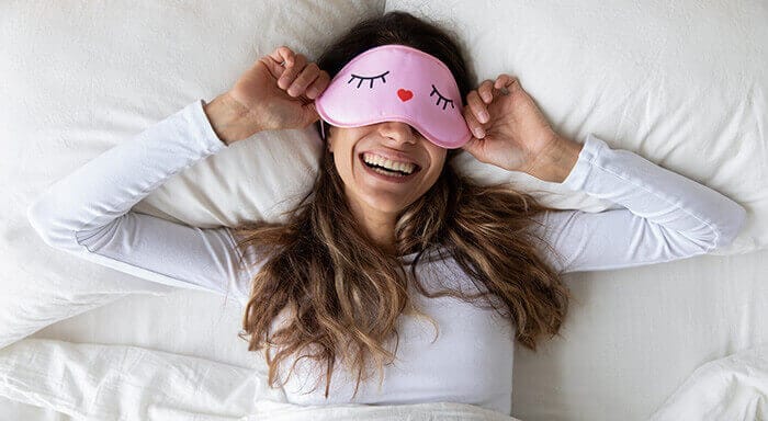 femme souriante dans son lit portant un masque de nuit aux yeux rieurs